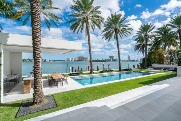 A contemporary waterfront residence viewed from its landscaped pool terrace. The image features a long rectangular swimming pool, expansive pale stone or concrete paving, manicured lawn, tall palm trees, and a minimalist white flat-roof pavilion with an outdoor dining area and kitchen visible beneath the overhang. A broad body of water and distant high-rise buildings form the background.