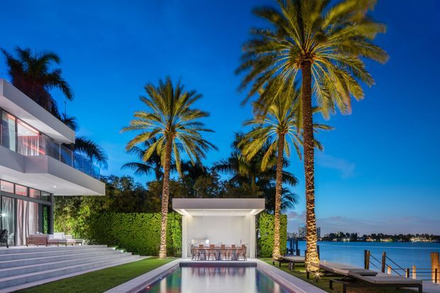 A luxurious modern waterfront residence at dusk, featuring crisp white geometric architecture, expansive glass walls, broad stepped terraces, a landscaped lawn, tall illuminated palm trees, and a narrow swimming pool aligned toward the calm water beyond. A covered outdoor dining pavilion sits beside the pool, while a private dock and distant shoreline are visible in the background.