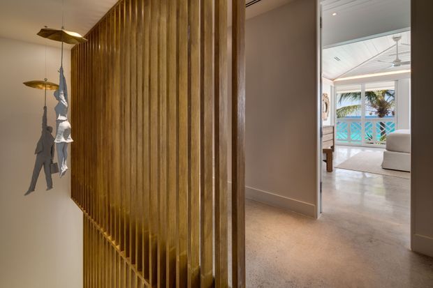 Interior view of a refined hallway or entry passage featuring a tall vertical timber slat screen with integrated stair detailing. Warm pendant lights and a sculptural hanging element add interest, while an open doorway leads into a bright living area with coastal views, a balcony railing, and a ceiling fan.