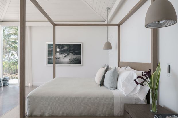 A bright, serene bedroom with a neatly made bed, pale textured bedding, white walls, and warm timber framing. The room features a pitched white-lined ceiling with exposed timber beams, pendant lights, a dark timber bedside surface, and a tall vase of tropical foliage. Glazing or an open connection at left reveals greenery and a pale outdoor area.