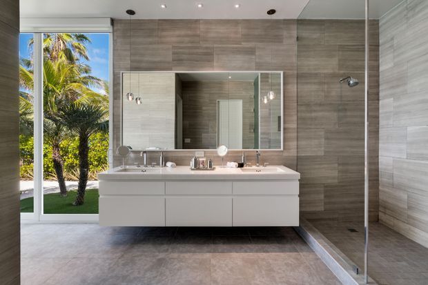 Contemporary bathroom with a floating double vanity, large mirrored cabinet, walk-in glass shower, and full-height grey wood-look tile surfaces. A full-height glazed door or window opens to a tropical garden with palm trees, bringing strong natural light into the space.