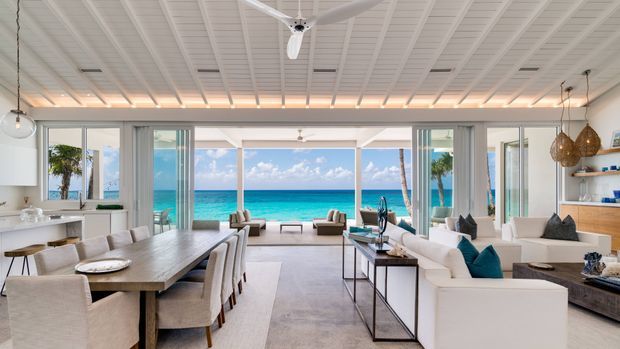 Bright open-plan coastal interior with a dining area, kitchen, and living room arranged around expansive glazed doors framing a turquoise ocean view. The room features white-painted ribbed ceiling panels, light neutral furnishings, a large timber dining table, pale flooring, woven pendant lights, and turquoise accent cushions. The architecture emphasizes indoor-outdoor connection and beachfront outlook.