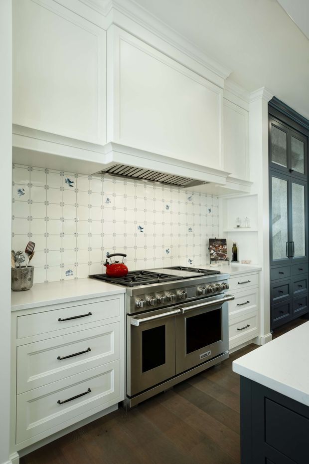 A refined traditional kitchen featuring white shaker-style cabinetry with decorative panel moulding, a large stainless-steel range cooker, patterned white tiled splashback, dark timber flooring, and a contrasting dark built-in cabinet wall. The space has detailed crown moulding, a concealed rangehood, black hardware, and a light benchtop, creating a classic high-end look.