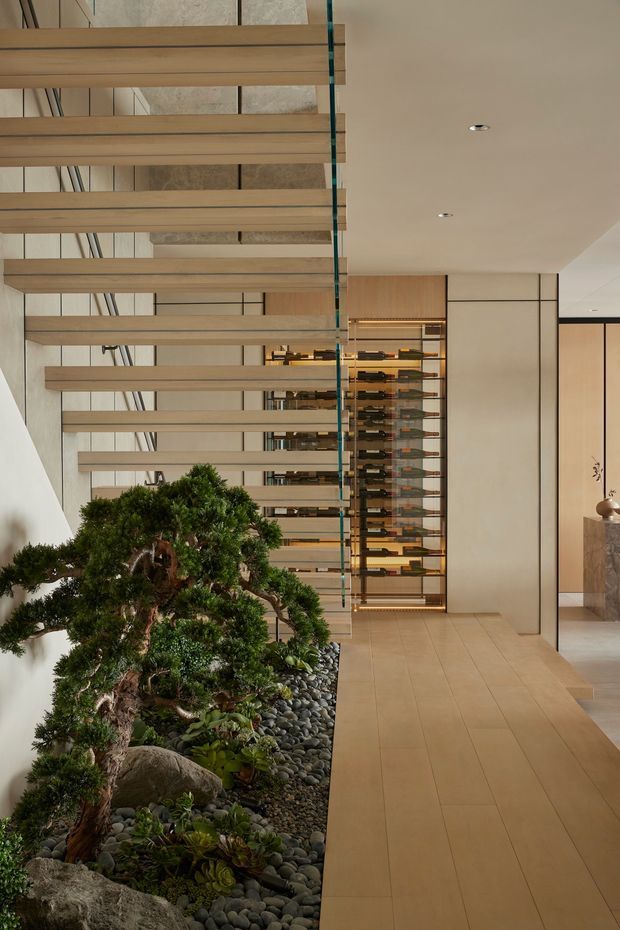 A refined contemporary interior hallway or landing featuring a sculptural open-riser timber staircase with glass balustrade, a tall built-in wine display, pale stone-look wall panels, and warm wood cabinetry. A carefully landscaped indoor Japanese-inspired garden with bonsai-like trees, rocks, succulents, and dark river pebbles runs alongside the circulation space. The palette is warm neutral and minimalist, with concealed and recessed lighting.
