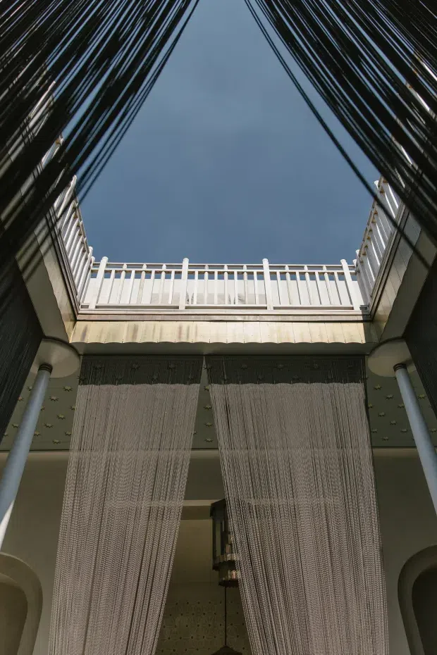 Vertical view through a dramatic interior courtyard or atrium, framed by dark reflective walls and pale columns. A white balustrade and upper-level balcony surround an open section of blue sky, while long textured sheer curtains hang down into the space. Decorative patterned wall surfaces and an ornate hanging light are visible below, creating a refined, theatrical atmosphere.