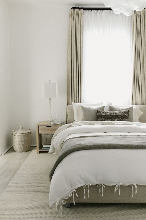 A softly styled bedroom with a large upholstered bed, layered white and grey bedding, neutral curtains framing a bright window, a timber bedside table with lamp, woven basket, and a textured carpet. The palette is calm and understated, with subtle natural-texture accents and a cloud-like ceiling pendant.