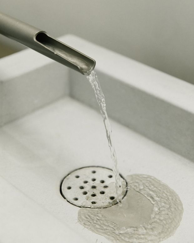 Close-up detail of water flowing from a brushed metal spout into a white rectangular basin or trough, passing over a circular perforated drain cover. The image is tightly framed around the plumbing fixture and drainage area, with a neutral, minimalist appearance.