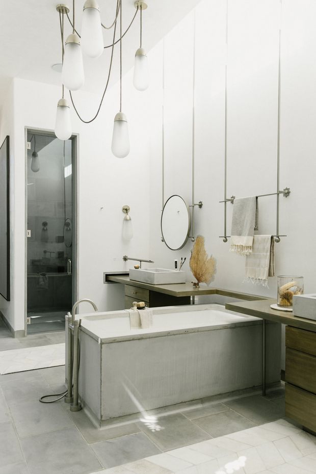 A bright, sculptural contemporary bathroom with a freestanding rectangular bathtub in the foreground, a floating double vanity with pale stone basins, wall-mounted taps, a large round mirror, and open industrial-style towel rails. The room has very high white walls, grey tiled flooring, exposed plumbing details, and multiple suspended white pendant lights. A glass-enclosed shower is visible to the left, with restrained neutral finishes and a few dried botanical accents.