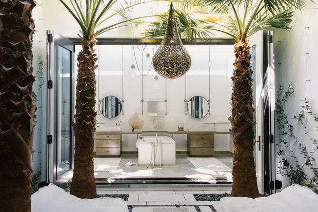 A luxurious, light-filled bathroom viewed through open glass doors, framed by two tall indoor palm trees. The space features a freestanding white bathtub, twin floating timber-toned vanity units, round mirrors, pale stone surfaces, and a large textured metallic pendant light. White walls, patterned floor inlays, and indoor greenery create a resort-like tropical atmosphere.