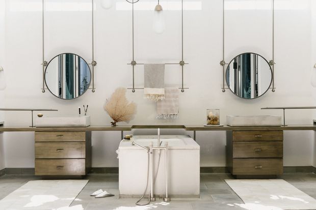 A refined, symmetrical bathroom vanity wall with two round mirrors, long floating or console-style cabinetry, twin drawer units in a weathered wood finish, and a freestanding rectangular bathtub centered in the foreground. The space uses pale plaster walls, light stone or concrete-look flooring, white cabinetry, brass-toned mirror hardware, and soft neutral styling. Large translucent or glazed wall panels create a bright, serene atmosphere.
