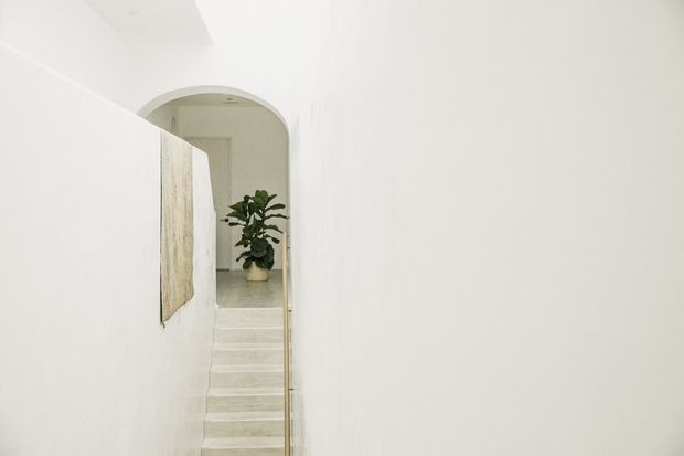 Minimal, softly lit interior view of a narrow stairway with pale stone or concrete steps, white plaster walls, and a rounded arched opening at the upper landing. A slender brass-toned handrail runs alongside the stairs, with a potted plant and understated wall art adding subtle detail.
