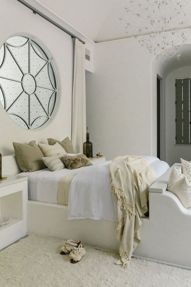 A bright, refined bedroom with a built-in platform bed dressed in layered white and beige bedding. The room features a large circular decorative wall panel with geometric detailing, a curved arched doorway, pale walls, a plush light carpet, integrated bedside joinery, and a sculptural branching pendant light. Neutral furnishings and soft textiles create a calm, luxurious atmosphere.