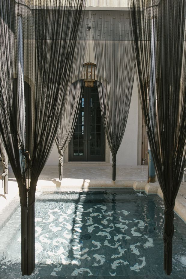 A dramatic indoor courtyard or pool pavilion with a small rectangular plunge pool in the foreground. Floor-to-ceiling dark-framed glazing and pale plaster walls sit behind a dense curtain-like installation of black and grey vertical cords, gathered around slender supports. A suspended lantern hangs centrally above the space, and sunlight creates strong reflections across the water and pale stone floor. The overall atmosphere is moody, luxurious, and highly textural.