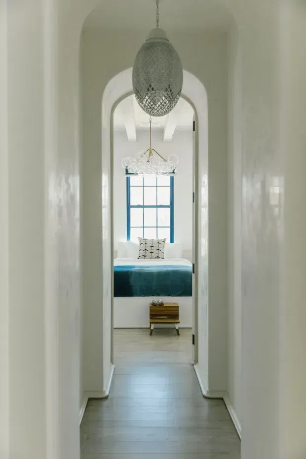 A narrow, bright hallway with white plastered walls and an arched opening leading into a bedroom. The bedroom contains a low bed with a vivid teal-blue cover, a patterned accent cushion, a blue-framed multi-pane window, a small timber bedside stool, and decorative pendant lighting. The hallway features pale timber-look flooring and a large woven pendant suspended centrally, with a second chandelier visible beyond. The composition emphasizes axial views, arched architecture, and layered interior spaces.