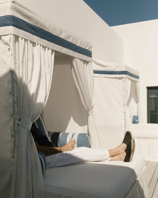 Sunlit outdoor cabana daybeds with white fabric curtains and canopy trim accented in muted blue. A person lounges on a cushioned daybed, with another matching cabana and a white architectural wall visible behind. The image has a relaxed, Mediterranean or resort-like atmosphere.