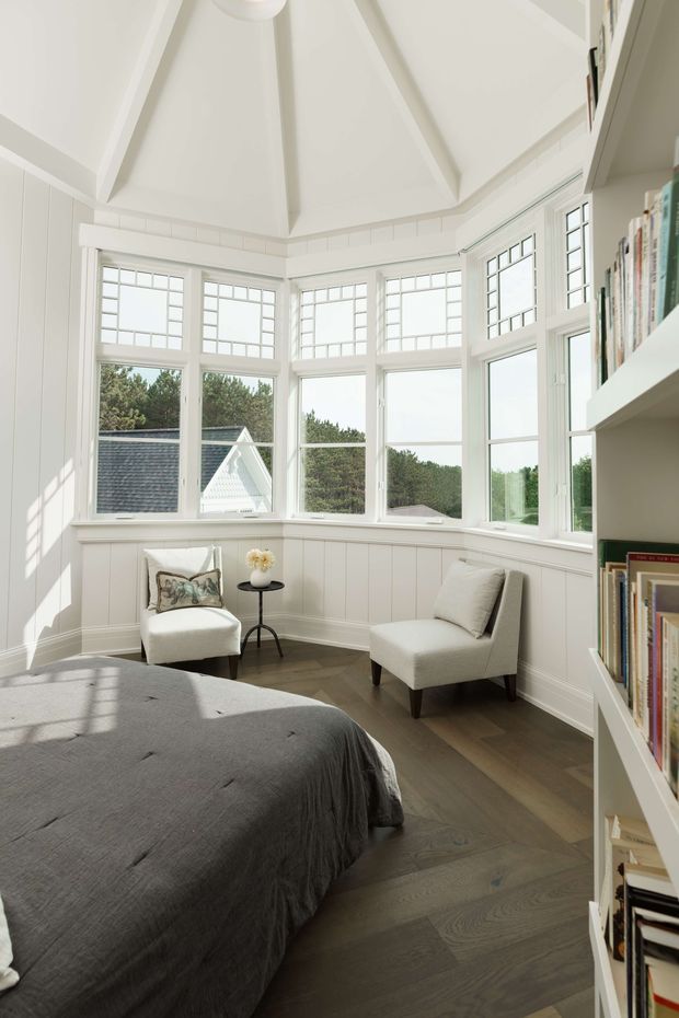 Bright, serene bedroom with a dramatic vaulted ceiling and a bay of tall white multi-pane windows. White wall panelling and trim surround a dark timber floor, while a large upholstered bed, two light armchairs, a small round side table, and built-in white bookshelves create a refined reading-and-resting space. Daylight and garden views fill the room.