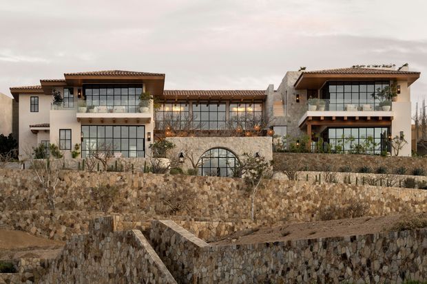 Wide exterior view of a large, multi-level residential building composed of several connected wings. The facade combines pale stucco, extensive dark-framed glazing, exposed timber balcony structures, terracotta-tiled low-pitched roofs, and substantial irregular natural-stone retaining walls. Landscaping and terraces are visible around the building, with a central arched glazed opening.