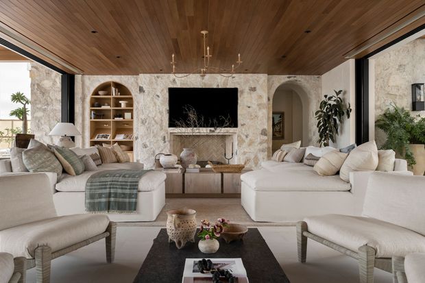 A refined, spacious living room with large cream sectional sofas and upholstered armchairs arranged around a dark rectangular coffee table. The room features a richly veined stone fireplace and television wall, built-in timber shelving, a warm timber-lined ceiling with recessed lighting, arched doorway details, and expansive black-framed glazing opening toward an outdoor setting. The palette is warm and neutral with layered natural textures and understated decorative furnishings.