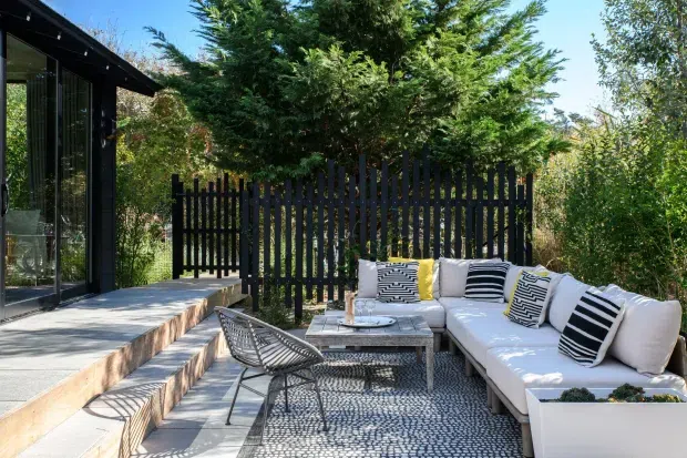 A bright, landscaped outdoor lounge beside a modern glass-walled structure. The space features a large white sectional sofa with black-and-white patterned cushions, a low weathered timber coffee table, a woven wire lounge chair, and a patterned outdoor rug on a paved terrace. Timber steps lead to the glazed building, while a tall black vertical-slat privacy fence and dense greenery enclose the area.