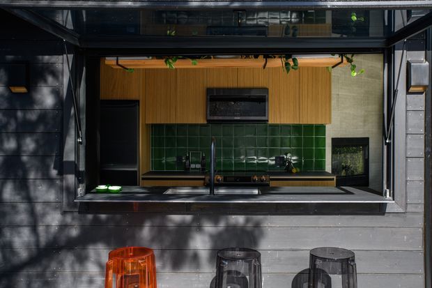 A compact outdoor-facing kitchen servery set into a dark horizontal-clad facade. The opening has a deep black-framed awning or counter window, with a stainless-steel sink and tap, dark benchtop, green glazed tile splashback, and warm timber cabinetry. Several stools sit outside beneath the serving opening, while plants and strong dappled sunlight add a relaxed residential character.