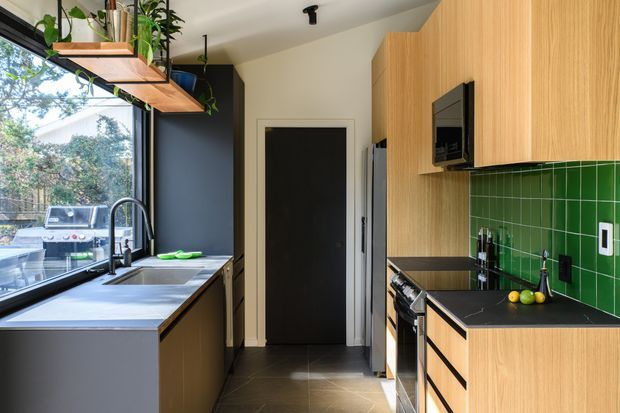 A narrow, contemporary galley kitchen with light timber cabinetry, dark benchtops, and a vivid green tiled splashback. The room features a large window beside a stainless-steel sink, black tapware, integrated appliances, open timber shelving with trailing plants, and dark floor tiles. A rear doorway and full-height cabinetry emphasize the compact linear layout.