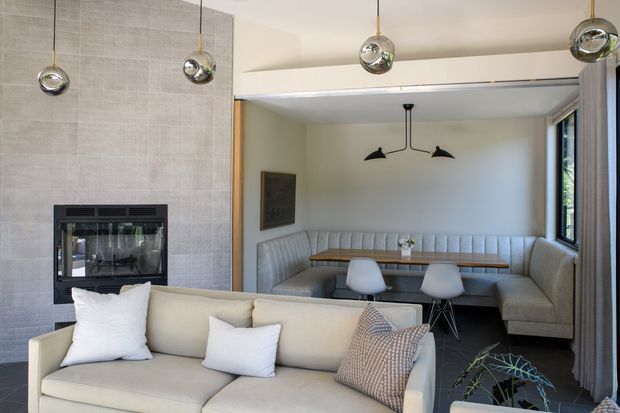 Bright contemporary open-plan living and dining area with a light neutral palette. A large cream sectional sofa faces a tiled fireplace wall, while a built-in upholstered banquette wraps around a timber dining table in the adjoining dining zone. Sculptural pendant lights, dark window frames, and restrained furnishings create a refined, comfortable interior.