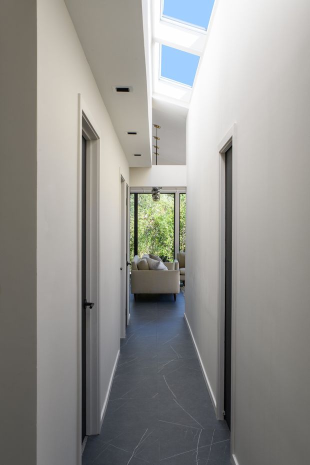 A bright, narrow interior hallway with white walls, multiple doorways, dark grey tiled flooring, recessed downlights, and a tall skylight above. The corridor opens toward a living room with a light sofa and full-height glazing overlooking greenery.