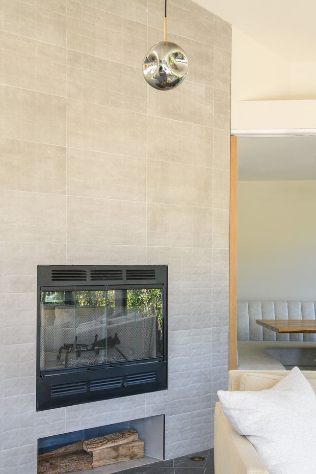 Interior view of a contemporary living room centred on a tall tiled fireplace wall with a black built-in firebox and log storage below. A polished chrome spherical pendant hangs in front of the wall, while a cream sofa and a built-in dining nook are visible to the right through an opening or reflection. The space has light neutral finishes, strong vertical proportions and abundant natural light.