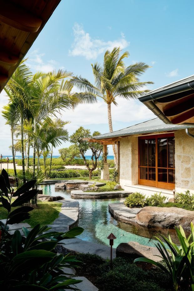 A tropical resort-style courtyard garden beside a low-rise building, featuring a narrow landscaped water feature with stepping stones, lush palms and broad-leaf planting, natural stone walls and paving, and timber-framed glazing. The building has a low-pitched metal roof with broad eaves and textured pale stone walls. Bright blue sky and a glimpse of ocean are visible beyond the garden.