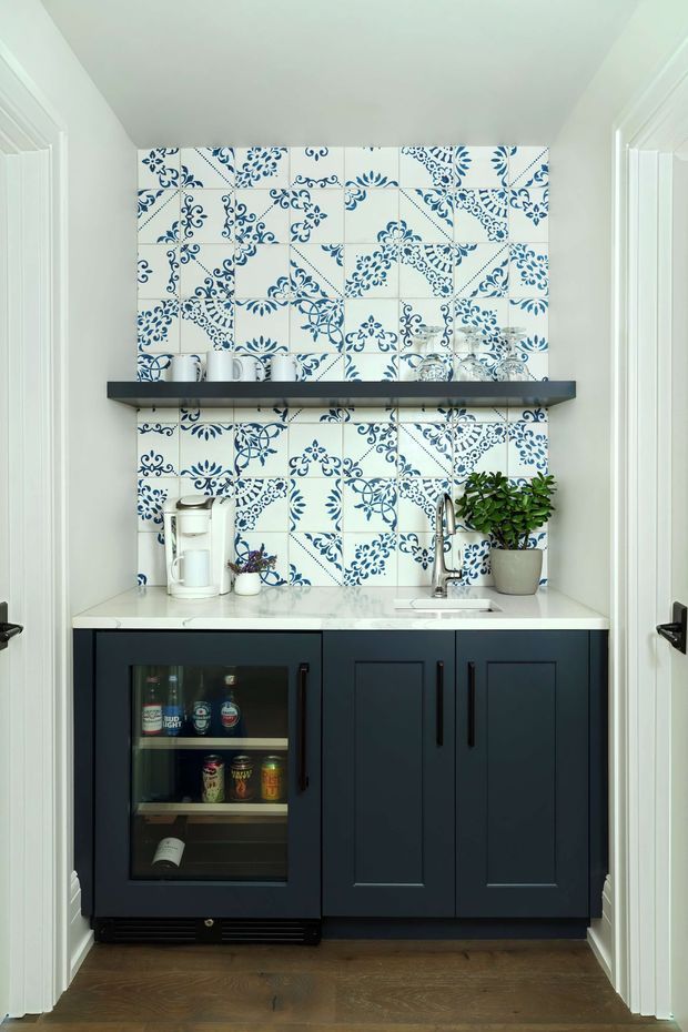 A compact built-in home bar or beverage station set within a small alcove. The space features deep navy-blue shaker cabinetry, a white engineered-stone benchtop, a glass-front undercounter drinks fridge, a stainless-steel sink and tap, and open dark shelving. The rear wall is covered in decorative white ceramic tiles with intricate blue floral and scroll patterns. White painted trim and walls frame the alcove, while dark timber flooring adds warmth.