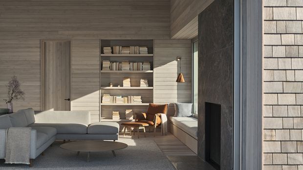A warm, minimalist living room with horizontal timber wall cladding, built-in open shelving, a large grey sectional sofa, lounge chairs, a low coffee table, and a dark stone fireplace surround. Natural light enters from the right, where a textured masonry-clad exterior wall is visible beyond the glazing. The space has a calm, refined contemporary cabin-like character with strong timber and stone finishes.