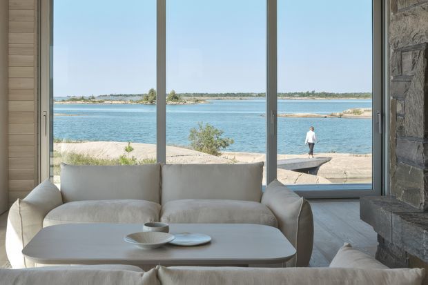 A calm, light-filled living room overlooking a body of water through expansive floor-to-ceiling windows. The room features a pale upholstered sofa, low rounded rectangular coffee table, light timber flooring and wall cladding, and a substantial textured stone fireplace wall. The restrained neutral palette and strong connection to the waterfront create a serene contemporary interior.