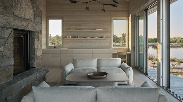 A refined contemporary living room with a large stone fireplace, pale timber wall panelling, built-in cabinetry and open shelving, and a soft neutral sofa arrangement. Full-height glazed sliding doors open to a waterside terrace, bringing in abundant natural light and views of the surrounding landscape. A dark sculptural ceiling light and restrained furnishings create a calm, modern atmosphere.