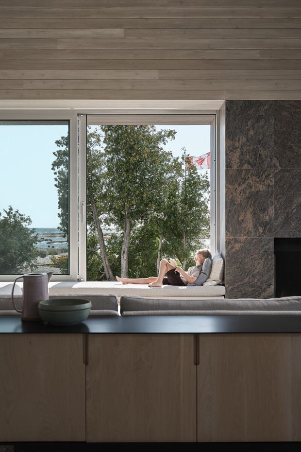 A refined contemporary living room with a built-in window seat overlooking a coastal waterfront. Warm horizontal timber wall panelling continues across the ceiling, while a dark natural-stone fireplace surround contrasts with pale upholstered seating and a dark benchtop or console in the foreground. Large windows provide abundant natural light and frame trees, water and a distant flag outside.