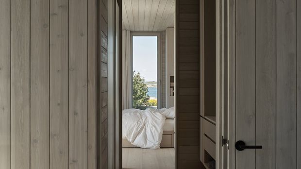 A narrow timber-lined passage or entry space leading into a compact bedroom. Pale vertical timber wall cladding and ceiling boards contrast with darker timber joinery and a black lever handle. The bedroom contains an unmade bed and opens toward a large glazed doorway or window with a distant coastal-looking view.