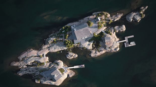 Aerial view of a group of modern buildings and decks integrated into rocky islands surrounded by dark water. The site includes flat-roofed structures, stone outcrops, planted areas, and several projecting piers or platforms.