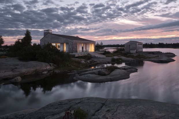 A low, dark-clad house and separate outbuilding sit among smooth exposed rock and calm water at dusk. Warm light glows through the large windows, contrasting with the cool purple-grey sky and rugged shoreline. The scene has a remote, minimalist coastal character.
