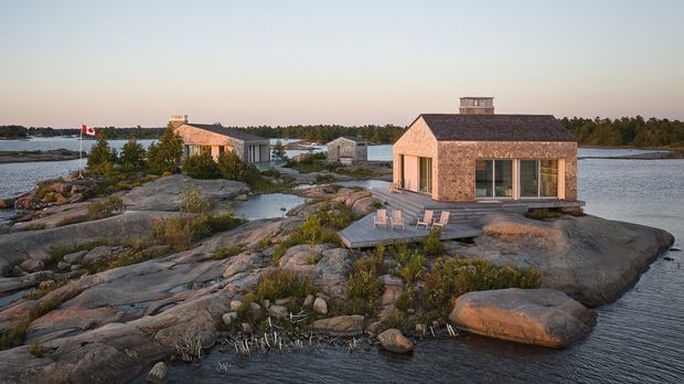 A small timber-clad waterfront dwelling on a rocky island, with a dark pitched roof, broad glazed openings, and a timber deck with outdoor chairs. Additional compact buildings sit behind it among exposed granite rocks and low coastal vegetation. The scene is photographed at warm evening light and includes a visible Canadian flag.