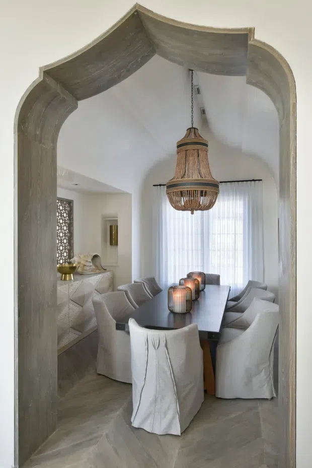 A refined dining room viewed through a large ornate arched doorway. The room features a dark rectangular dining table surrounded by light slipcovered chairs, a large woven pendant chandelier, sheer curtains over a tall window, white sculptural walls, and a textured light-wood sideboard. The floor and doorway are finished in weathered grey timber tones, creating a warm, elegant, contemporary interior.