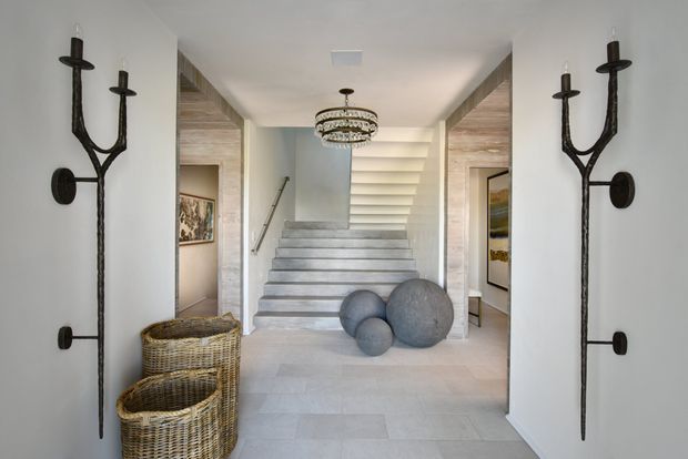 A refined residential entry hall viewed toward a broad staircase. The space features pale stone or tile flooring, light-toned walls, timber-lined recessed openings, a decorative crystal chandelier, sculptural dark metal wall sconces, woven basket storage, framed artwork, and large round stone-like decorative objects at the foot of the stairs. The staircase rises to a bright upper landing, creating an airy and elegant composition.