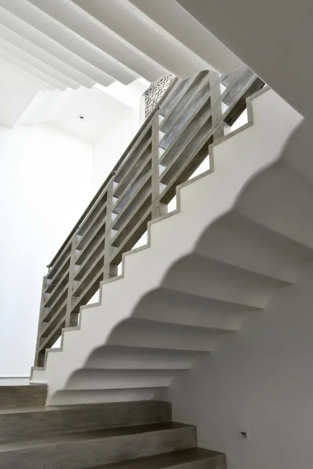 Interior view of a contemporary stairway with broad white sculptural stair stringers and risers, dark weathered timber treads, and a distinctive open timber balustrade with horizontal slats. White walls and a white ribbed or beam-like ceiling create a bright, high-contrast minimalist composition.
