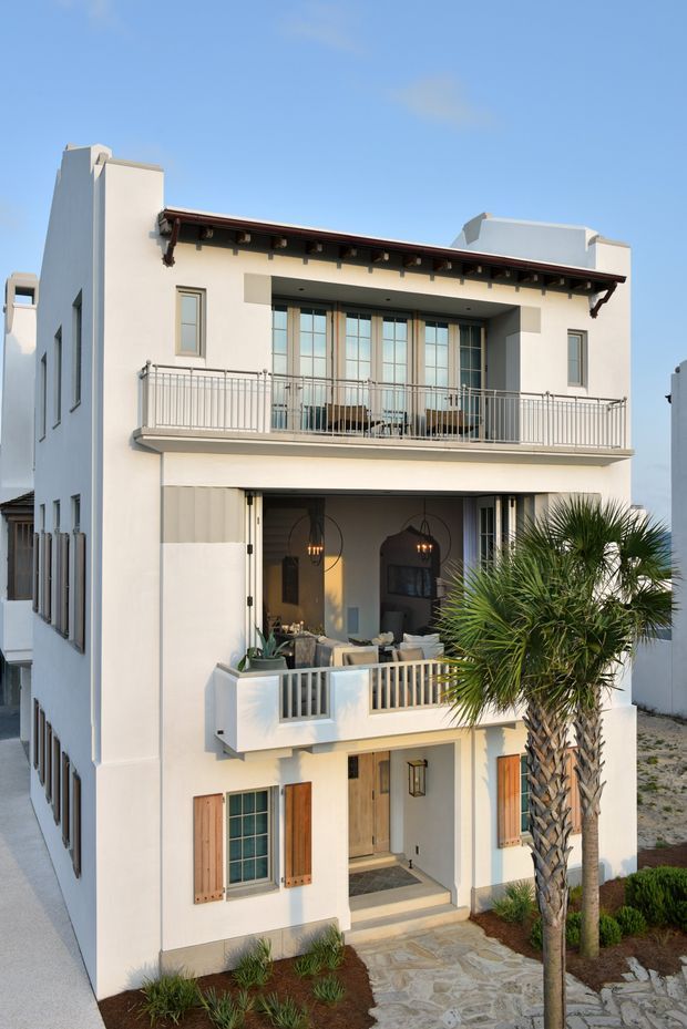 Front exterior view of a tall, narrow multi-storey residence with a light stucco facade, projecting balconies, timber shutters, large gridded windows, exposed dark roof eaves, and a recessed outdoor living area. A palm tree and landscaped stone entry path frame the facade.