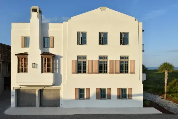 Front exterior view of a large, pale stucco multi-storey residence with a symmetrical facade, tall multi-pane windows, timber shutters, projecting bay windows, and a prominent chimney. The building sits beside a paved driveway and landscaped grounds under clear daylight.