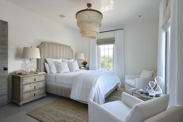 Bright, softly styled bedroom with a large upholstered bed, layered white bedding, matching bedside furniture, armchairs, and a round glass-topped side table. The room features white walls and curtains, a tall window with a Roman blind, a textured neutral rug, and an ornate tiered pendant light, creating a refined, calm and airy atmosphere.