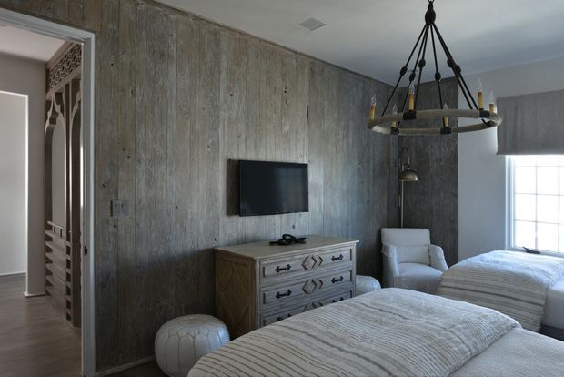 A calm, refined bedroom with two beds, upholstered white bedding, a light wood dresser, a wall-mounted television, and a white armchair. The room features a dramatic textured grey-brown vertical wall finish, a dark metal chandelier, and a large gridded window providing natural light. An adjacent opening reveals decorative architectural screening and a hallway beyond.