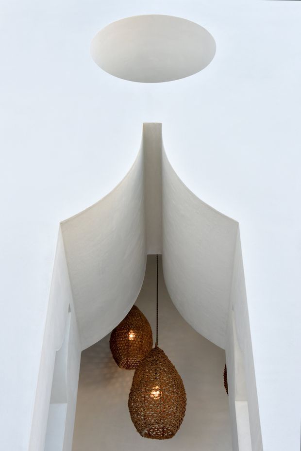 A minimalist interior architectural detail viewed upward through a tall white plastered opening or stairwell. Sculptural curved wall forms create a vaulted, flowing enclosure, with a circular ceiling opening above. Two woven natural-fibre pendant lights hang within the void, adding warm texture against the white surfaces.