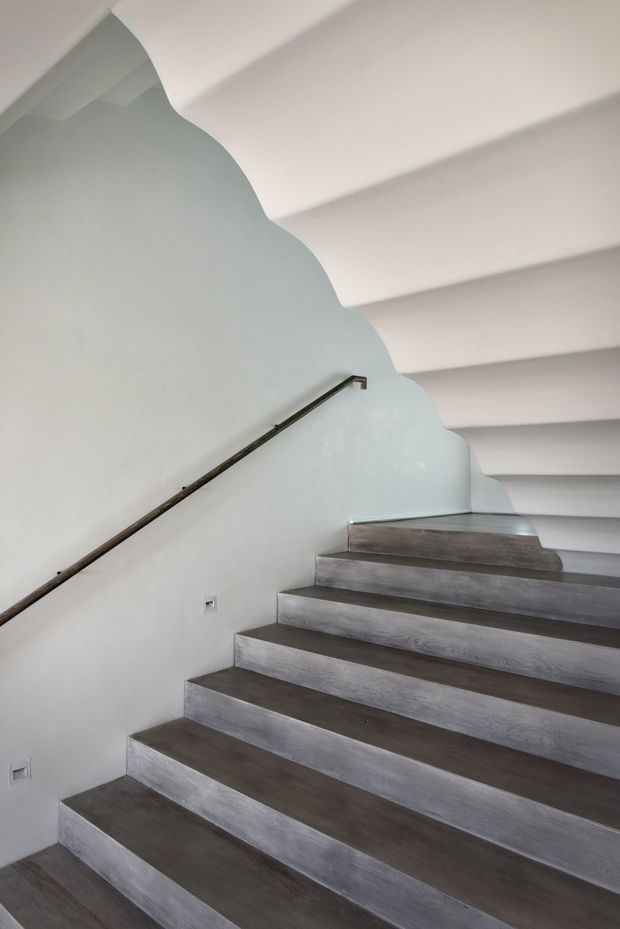 Minimalist interior stairway with broad dark timber-toned treads, pale smooth walls, a slim dark metal handrail, and a distinctive undulating scalloped wall edge beneath a white stepped or ribbed ceiling.
