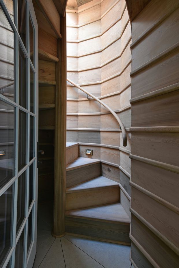 A compact curved stairway enclosed by richly detailed timber panelling. The staircase has wedge-shaped wooden treads, a smooth sculptural handrail, and a narrow window providing natural light. A glazed, multi-pane door and dark timber shelving are visible at the left edge. The space has a warm, handcrafted, highly tactile character.