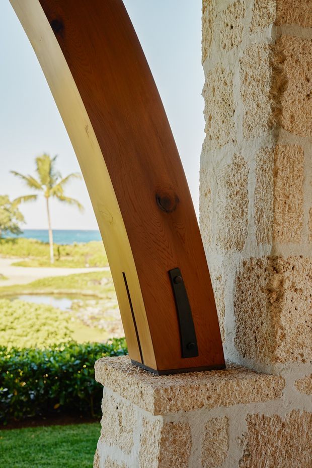 Close-up architectural detail of a large angled timber structural beam anchored to a textured pale stone masonry column. The beam has a warm dark-wood finish with visible grain and a black metal fixing plate. Through the opening, a tropical coastal landscape with palms, lawn, and ocean is visible.