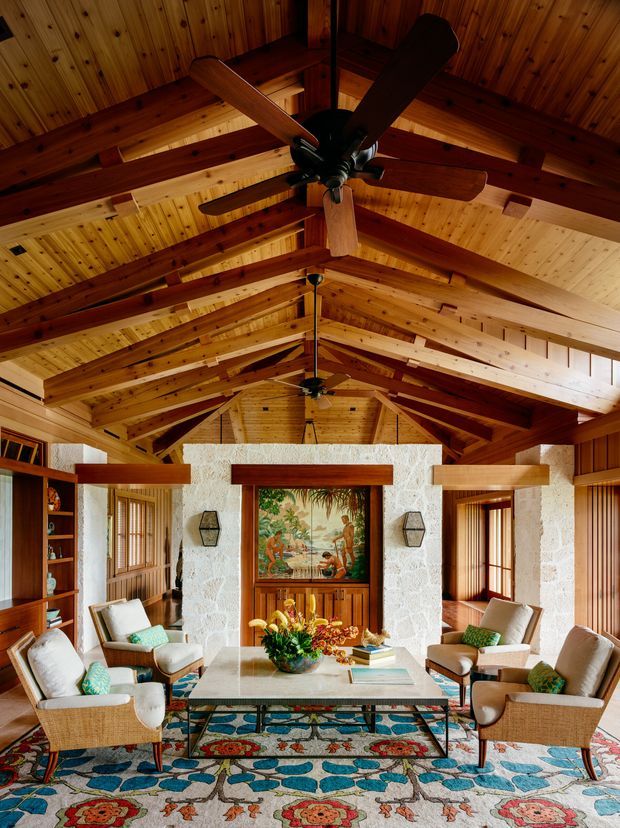 A warm, richly detailed living room with a dramatic exposed timber vaulted ceiling, heavy timber rafters and beams, and multiple ceiling fans. The room features pale textured stone walls, timber-framed windows and doors, built-in shelving, four light upholstered lounge chairs, a central stone-topped coffee table, and a colourful patterned rug. A decorative artwork or screen forms a focal point on the rear wall.
